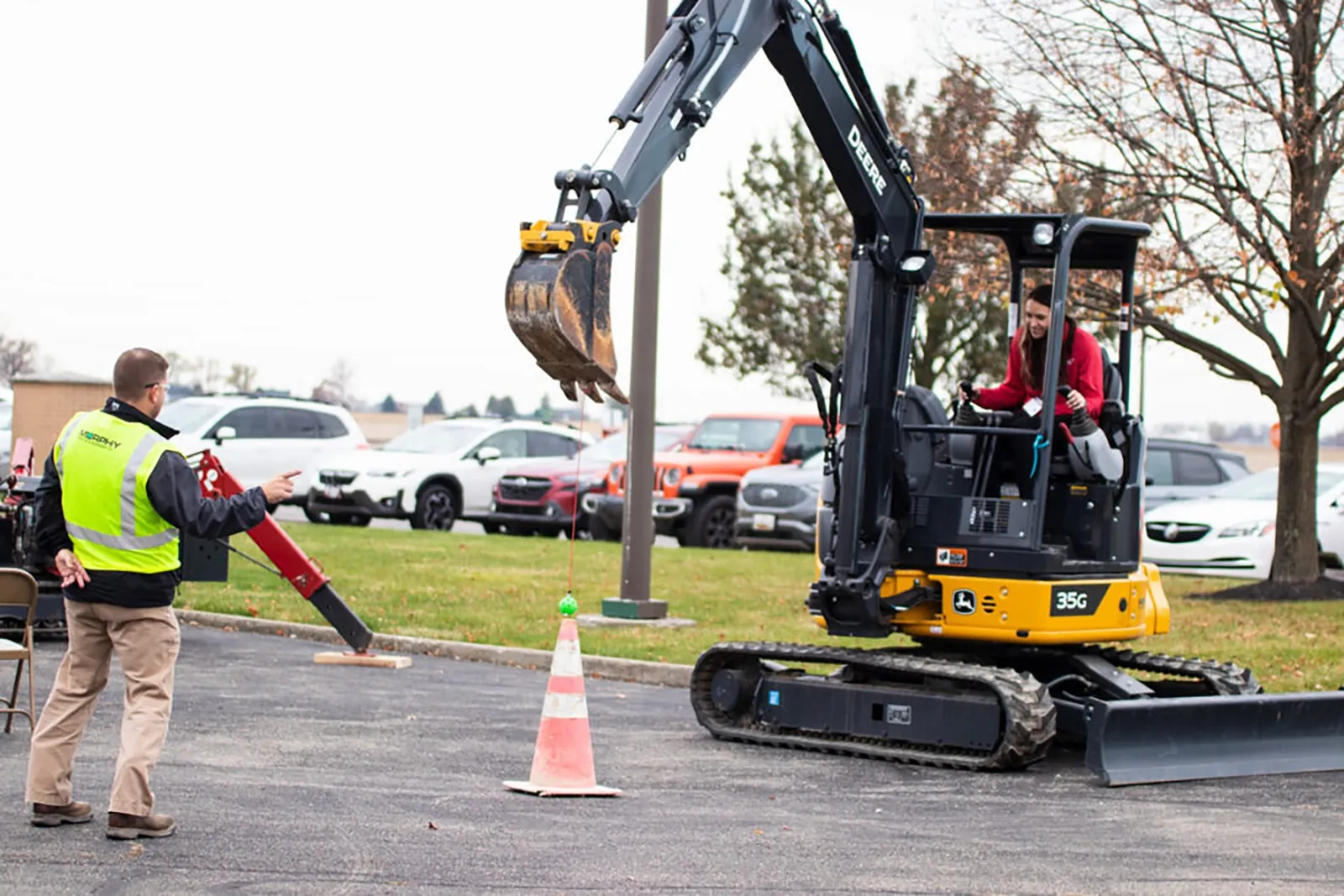 A person operates a compact excavator while another person in a safety vest stands nearby